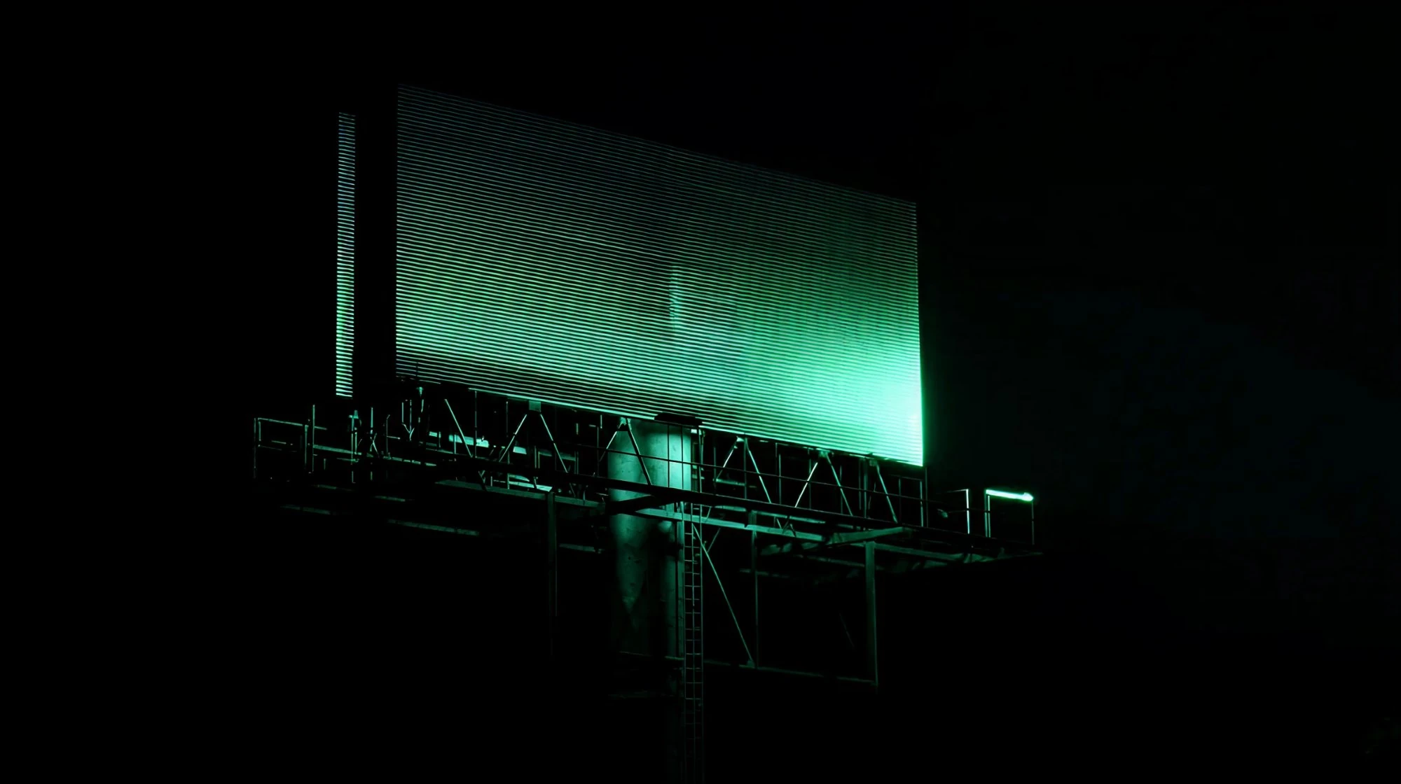 Large digital billboard glowing with green light at night, showing its metal support structure silhouetted against a dark sky. Offline signage tracking