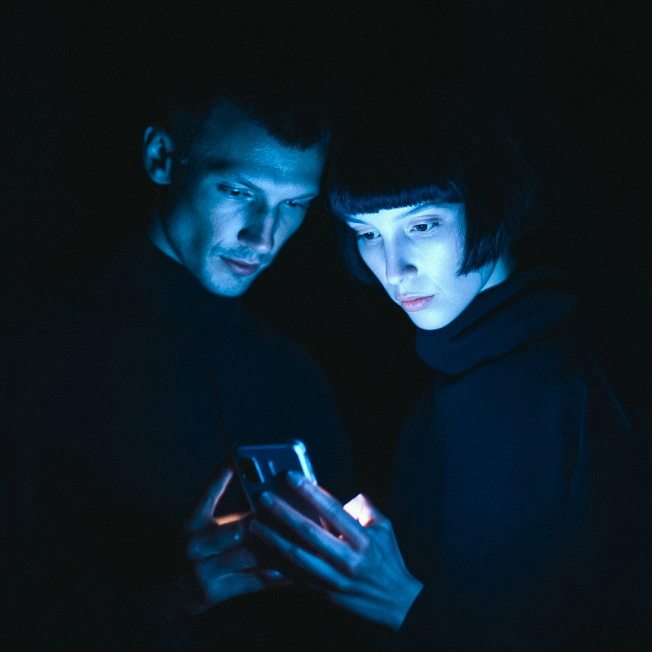 Two people in a dark room illuminated only by the blue glow of a smartphone screen they're both looking at intently, creating a moody atmospheric scene that evokes themes of digital connection and social media engagement.
